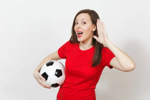 Beautiful European young woman, football fan or player in red uniform eavesdrop, hearing gesture, hold soccer ball isolated on white background. Sport play football, health, healthy lifestyle concept.