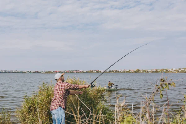 Sidan Visa ung orakad man med fiskespö i rutig skjorta, keps och solglasögon kastar fiske pole på sjön från stranden nära buskar och vass. Livsstil, rekreation, fisherman fritid koncept. — Stockfoto