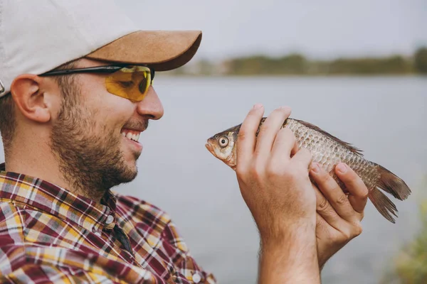 Close-up van jonge ongeschoren Glimlachende man in geruite shirt, pet en zonnebril een vis gevangen en kijkt naar het aan de oever van lake op achtergrond van water. Lifestyle, recreatie, visser leisure concept — Stockfoto