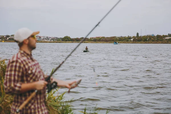 Young unshaven man in checkered shirt, cap, sunglasses pulled out fishing pole and holds caught fish on shore of lake near reeds on background of boat. Lifestyle, recreation, fisherman leisure concept
