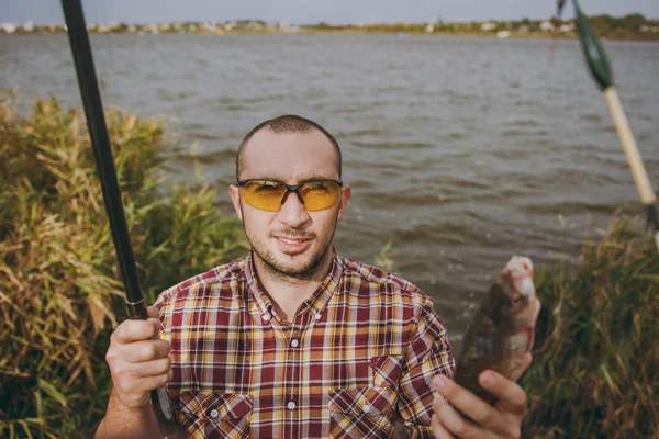 Joven hombre sonriente sin afeitar en camisa a cuadros y gafas de sol sacó poste de pesca y sostiene pescado capturado en la orilla del lago cerca de arbustos y cañas. Estilo de vida, recreación, concepto de ocio del pescador —  Fotos de Stock