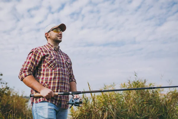 Jovem homem sem barba em camisa xadrez, boné e óculos de sol olhando em linha reta detém pólo de pesca e desenrolar carretel no céu de fundo, arbustos e juncos. Estilo de vida, recreação, conceito de lazer do pescador . — Fotografia de Stock