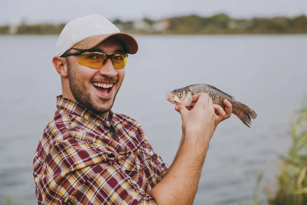 Close up Young unshaven smiling man in checkered shirt, cap and sunglasses caught a fish, holds it in arms and rejoices on shore of lake on background of water. Lifestyle, fisherman leisure concept — Stock Photo, Image