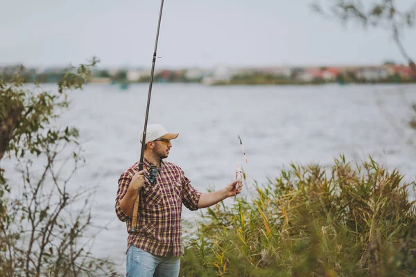 Sidan Visa ung orakad man i rutig skjorta, mössa, solglasögon drog ut metspö och innehar fångad fisk på stranden av sjön nära buskar och vass. Livsstil, rekreation, fisherman fritid koncept — Stockfoto