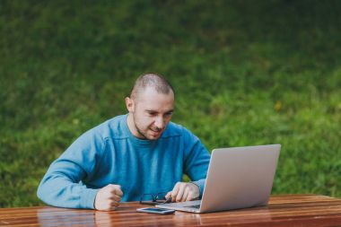 Genç üzgün kızgın şok adam işadamı veya öğrenci Şehir Parkı doğa açık havada sorunlar hakkında ilgili çalışan dizüstü kullanarak masada oturan rahat mavi gömlekli gözlüklü. Mobil ofis kavramı.