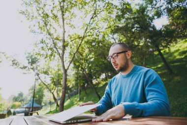 Genç başarılı akıllı adam işadamı veya öğrenci şehir Park açık havada yeşil doğa üzerinde çalışan dizüstü kullanarak cep telefonu ile masada oturan rahat mavi gömlekli gözlüklü. Mobil ofis kavramı.