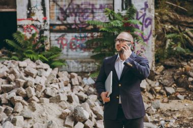 Beyaz gömlek, Klasik takım elbise, gözlük genç başarılı iş adamı. Dizüstü pc bilgisayar ile ayakta, Harabeleri, enkaz, taş açık havada binanın yakınındaki telefonda konuşan adam. Mobil ofis, iş kavramı.