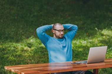 Genç işadamı veya öğrenci rahat mavi gömlekli, rahatlatıcı, tablo dizüstü bilgisayar ile hareket eden telefon kafa, arkasında elele Şehir Parkı içinde oturup gözlük açık havada çalışan adam. Mobil ofis kavramı.
