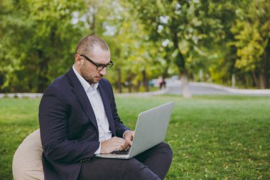 Beyaz gömlek, Klasik takım elbise, gözlük genç başarılı iş adamı. Adam dizüstü pc bilgisayar şehir Park açık havada doğa üzerinde yeşil çim üzerinde çalıştığı yumuşak kabarık üzerinde oturmak. Mobil ofis, iş kavramı.
