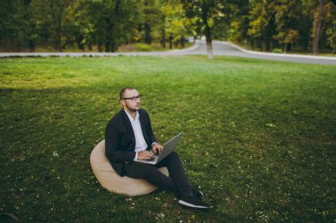 Beyaz gömlek, Klasik takım elbise, gözlük genç başarılı iş adamı. Adam dizüstü pc bilgisayar şehir Park açık havada doğa üzerinde yeşil çim üzerinde çalıştığı yumuşak kabarık üzerinde oturmak. Mobil ofis, iş kavramı.