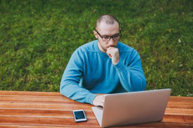 Genç dalgın başarılı akıllı adam işadamı ya da doğa dışarıda üzerinde çalışma öğrenci gözlük cep telefonu, dizüstü bilgisayar kullanarak şehir parkı ile masada oturan rahat mavi gömlekli. Mobil ofis kavramı.