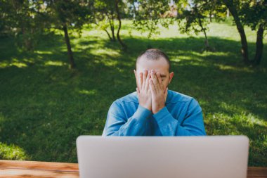 Genç üzgün adam işadamı yorgun veya açık havada, çalışan öğrenci rahat mavi gömlekli, cep telefonu, dizüstü bilgisayar kullanarak şehir parkı ile masada oturan gözlük sorunlar hakkında endişelendiriyor. Mobil ofis kavramı.