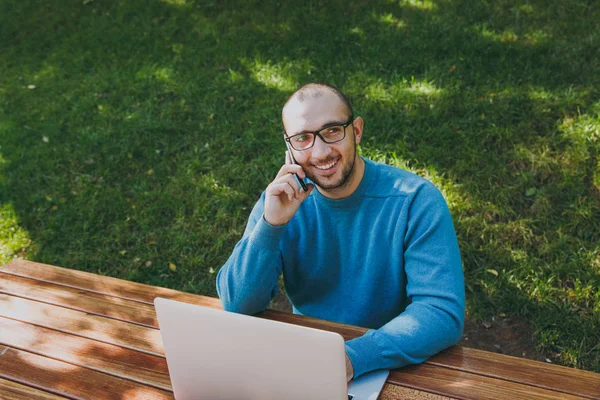 Young successful smiling smart man businessman or student in casual blue shirt, glasses sitting at table, talking on mobile phone in city park using laptop, working outdoors. Mobile Office concept.