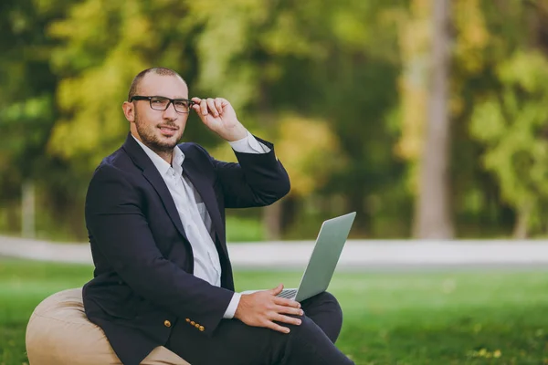 Successful businessman in white shirt, classic suit, correct glasses. Man sit on soft pouf, work on laptop pc computer in city park on green lawn outdoors on nature. Mobile Office, business concept.