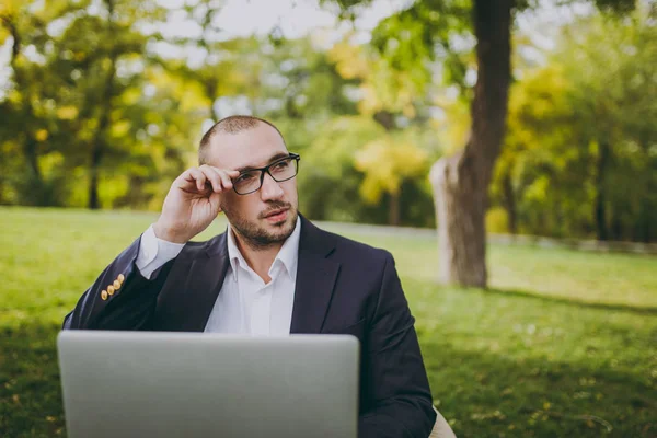 Молодой успешный бизнесмен в белой рубашке, классическом костюме, корректирует ручные очки. Мужчина сидит на мягком пуфе, работает с ноутбуком в городском парке на зеленой лужайке на открытом воздухе. Мобильный офис, концепция бизнеса . — стоковое фото