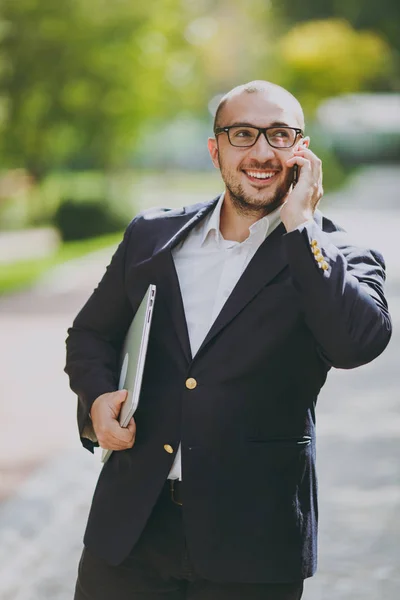 Empresário inteligente bem sucedido em camisa branca, terno clássico, óculos. Man stand com computador portátil pc, falar no telefone celular no parque da cidade ao ar livre no fundo da natureza. Escritório móvel, conceito de negócio . — Fotografia de Stock