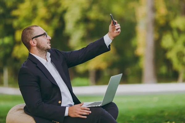 Молодой бизнесмен в белой рубашке, классическом костюме, очках. Человек сидит на мягком пуфе с ноутбуком компьютер, делая селфи на мобильный телефон в городском парке на зеленой лужайке на открытом воздухе. Концепция мобильного офиса . — стоковое фото