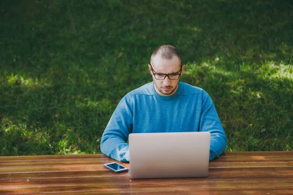 Giovane uomo intelligente di successo uomo d'affari o studente in casual occhiali camicia blu seduto a tavola con il telefono cellulare nel parco della città utilizzando computer portatile che lavora all'aperto su sfondo verde. Concetto Mobile Office . — Foto Stock