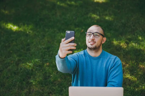 Młody udanej zabawy smart człowieka biznesmen lub student w casual niebieską koszulę, Okulary, siedząc przy stole robić selfie na telefon komórkowy w parku miejskim przy użyciu laptopa, pracy na zewnątrz. Koncepcja Mobile Office. — Zdjęcie stockowe