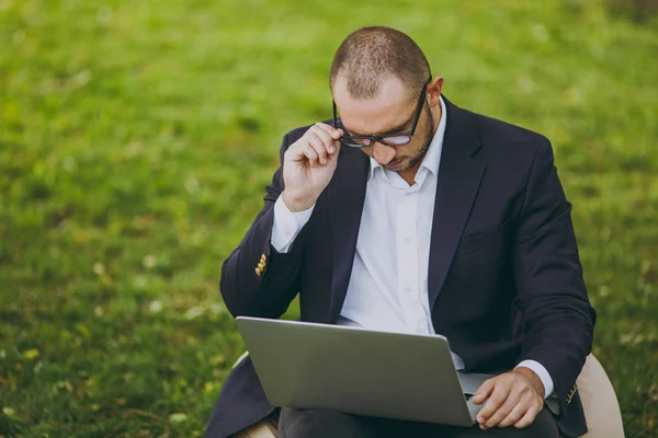 Успешный бизнесмен в белой рубашке, классический костюм, правильные очки. Человек сидит на мягком пуфе, работает на ноутбуке компьютера в городском парке на зеленой лужайке на природе. Мобильный офис, концепция бизнеса . — стоковое фото