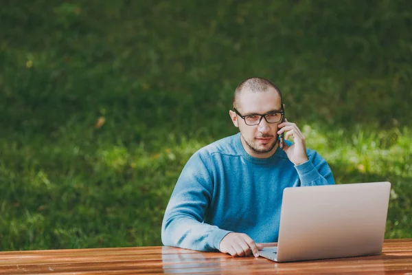 Młody udany uśmiechający się biznesmen człowiek inteligentny lub student w casual niebieską koszulę, Okulary, siedząc przy stole, rozmawiając na telefon komórkowy w parku miejskim przy użyciu laptopa, pracy na zewnątrz. Koncepcja Mobile Office. — Zdjęcie stockowe