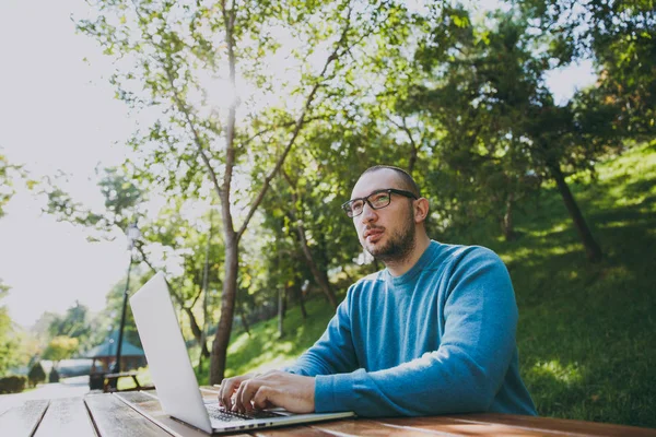Young successful smart man businessman or student in casual blue shirt glasses sitting at table with mobile phone in city park using laptop working outdoors on green nature. Mobile Office concept.