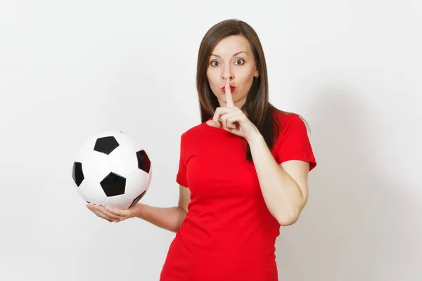 Young woman, football fan or player in red uniform hold soccer ball, say hush be quiet with finger on lips gesture isolated on white background. Sport, play football, cheer, healthy lifestyle concept.