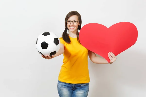 Mujer europea sonriente, divertidas colas de caballo, fanático del fútbol o jugador en gafas, uniforme amarillo sostienen la clásica pelota de fútbol, corazón rojo aislado sobre fondo blanco. Deporte, fútbol, concepto de estilo de vida saludable . —  Fotos de Stock