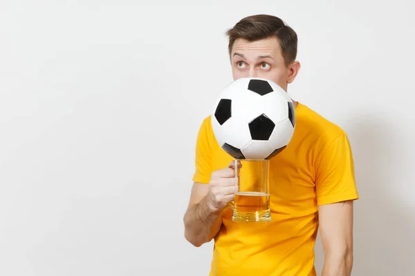 Joven divertido hombre europeo alegre, ventilador o jugador en uniforme amarillo se esconden detrás de jarra de cerveza, pelota de fútbol animar equipo de fútbol favorito aislado sobre fondo blanco. Deporte, juego, estilo de vida concepto . — Foto de Stock