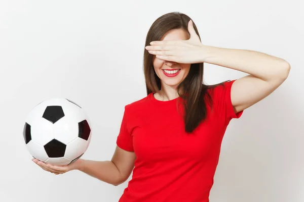 Beautiful European young woman, football fan or player in red uniform hold classic soccer ball, cover eyes by hand isolated on white background. Sport play football, health, healthy lifestyle concept.