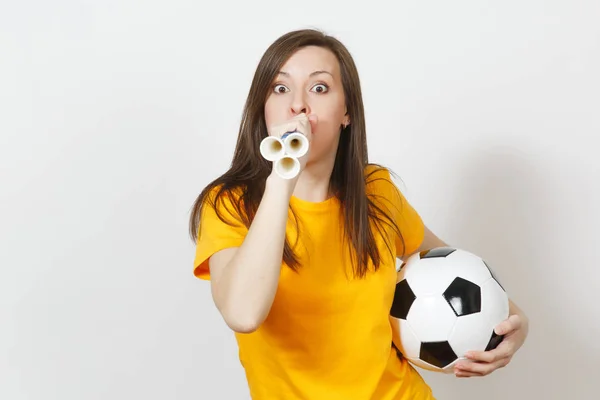 Beautiful European young cheerful happy woman, soccer fan or player in yellow uniform holds and blows football pipe, ball isolated on white background. Sport, play football, healthy lifestyle concept.