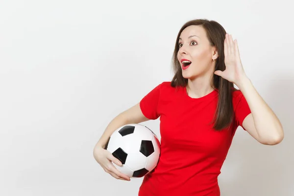 Beautiful European young woman, football fan or player in red uniform eavesdrop, hearing gesture, hold soccer ball isolated on white background. Sport play football, health, healthy lifestyle concept.