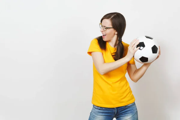 Fun cheerful European woman, two pony tails, football fan or player in glasses, yellow uniform hold classic soccer ball isolated on white background. Sport, play, football, healthy lifestyle concept.