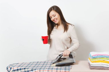 Kırmızı fincan çay veya kahve, Ütü gömlek, giyim demir ile Ütü Masası üzerine ışık giysili genç ev hanımı. Beyaz arka plan üzerinde izole kadın. Temizlik kavramı. Kopyalama alanı reklam.