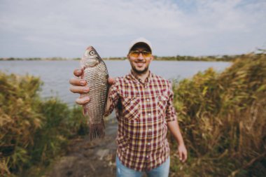 Tıraşsız delikanlı kareli gömlek, şapka ve güneş gözlüğü balık yakaladım, gülümser ve bu su, çalılar ve sazlık arka plan üzerinde gölün kıyısında gösterir. Yaşam tarzı, balıkçı dinlenme, eğlence kavramı