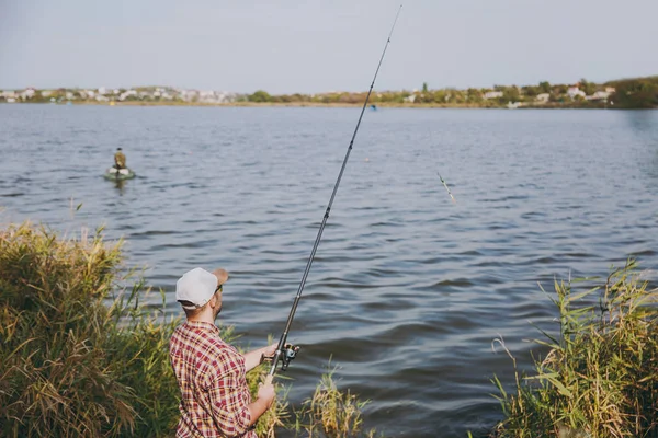 Vista posterior Joven hombre sin afeitar con caña de pescar en camisa a cuadros, gorra y gafas de sol lanza cebo y pesca en el lago desde la orilla cerca de arbustos y cañas. Estilo de vida, recreación, concepto de ocio del pescador . —  Fotos de Stock