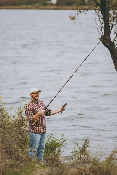 Ung orakad leende man i rutig skjorta, keps och solglasögon drog ut metspö och innehar fångad fisk på stranden av sjön nära buskar och vass. Livsstil, rekreation, fisherman fritid koncept — Stockfoto