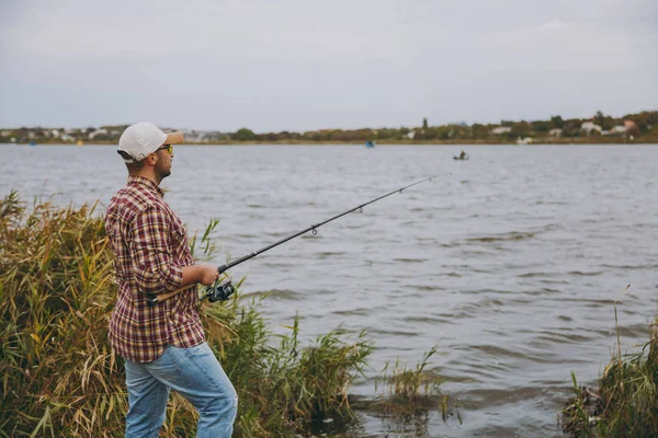 Vista lateral Joven hombre sin afeitar con una caña de pescar en camisa a cuadros, gorra y gafas de sol lanza caña de pescar en un lago de la orilla cerca de arbustos y cañas. Estilo de vida, recreación, concepto de ocio del pescador —  Fotos de Stock