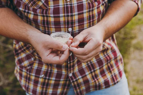 Cerrar pescador recortado en camisa a cuadros sostiene en las manos pequeña caja blanca con gusanos, poner cebo en el gancho para pescar con caña de pescar. El hombre tiene gusanos para pescar. Estilo de vida, recreación, concepto de ocio — Foto de Stock