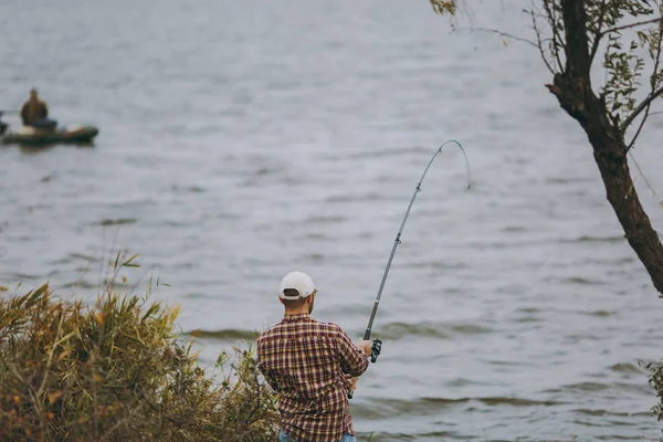 Ung man med ett fiskespö i rutig skjorta och keps kastar bete och fiske mot bakgrund av båt på sjön från stranden nära buskar och vass. Livsstil, rekreation, fisherman fritid koncept — Stockfoto