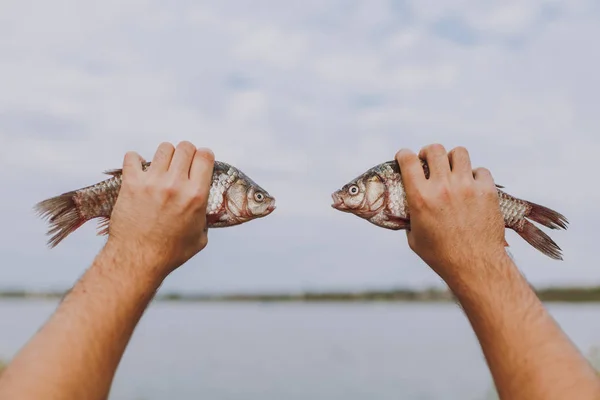 De cerca Un hombre sostiene en sus manos dos peces con la boca abierta uno frente al otro en un lago borroso y el fondo del cielo. Estilo de vida, recreación, concepto de ocio de los pescadores. Copiar espacio para publicidad . —  Fotos de Stock