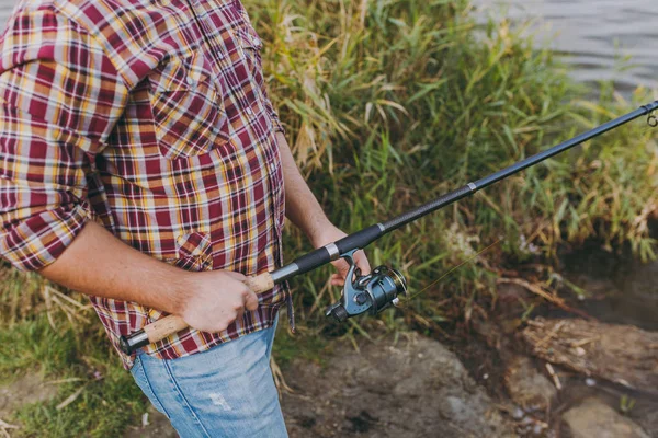 Un hombre con camisa a cuadros con mangas enrolladas sostiene un poste de pesca y desenrollando el carrete en la orilla del lago cerca de arbustos y cañas. Estilo de vida, recreación, concepto de ocio —  Fotos de Stock