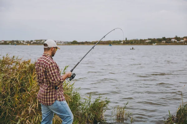 Visão lateral Jovem homem sem barba com uma vara de pesca em camisa quadriculada, boné e óculos de sol lança pólo de pesca em um lago da costa perto de arbustos e juncos. Estilo de vida, recreação, conceito de lazer do pescador — Fotografia de Stock