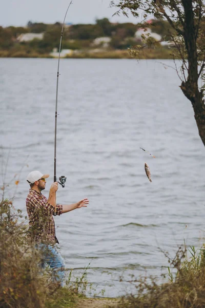 Vista lateral Joven hombre sin afeitar en camisa a cuadros, gorra y gafas de sol saca caña de pescar con pescado capturado en un lago de la orilla cerca de arbustos y cañas. Estilo de vida, recreación, concepto de ocio del pescador —  Fotos de Stock