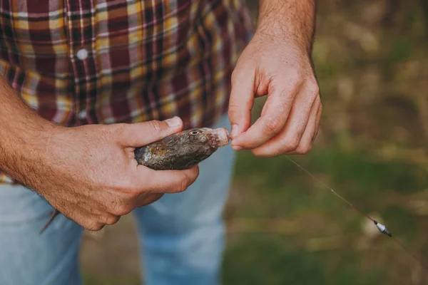 Close up Man in checkered shirt and jeans removes caught fish from a hook on fishing rod on a blurred green background. Lifestyle, recreation, fisherman leisure concept. Copy space for advertisement. — Stock Photo, Image