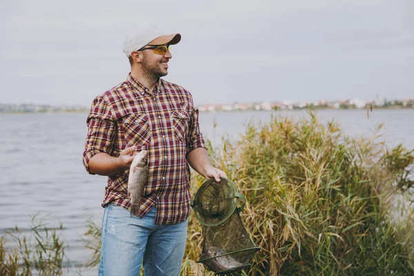 Joven hombre sonriente sin afeitar en camisa a cuadros, gorra, gafas de sol mirando hacia otro lado mantiene en las manos red de pesca verde y peces que capturó en la orilla del lago cerca de las cañas. Estilo de vida, concepto de ocio del pescador —  Fotos de Stock