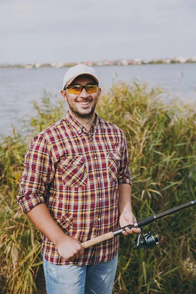 Joven hombre sonriente sin afeitar en camisa a cuadros, gorra y gafas de sol sostiene caña de pescar en la orilla del lago en el fondo de agua, arbustos y cañas. Estilo de vida, recreación, concepto de ocio del pescador . —  Fotos de Stock
