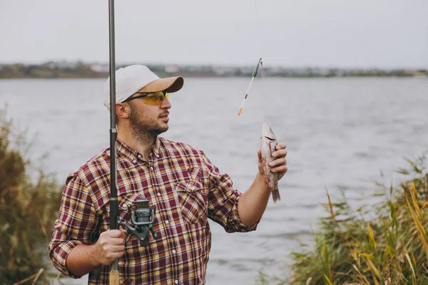 Jonge ongeschoren man in geruite shirt, pet en zonnebril trok een hengel en wachtruimten gevangen vis op de oever van het meer in de buurt van struiken en riet. Lifestyle, recreatie, visser leisure concept — Stockfoto