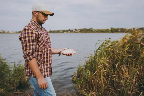 Il giovane uomo non rasato in camicia a scacchi, berretto e occhiali da sole ha preso un pesce e lo tiene in braccia su riva di lago su sfondo d'acqua, arbusti e canne. Stile di vita, ricreazione del pescatore, concetto di svago — Foto Stock