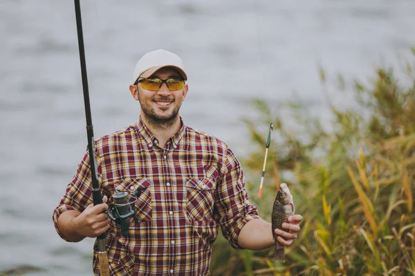 Giovane uomo non rasato in camicia a scacchi, berretto e occhiali da sole tirato fuori canna da pesca con il pesce catturato e gioisce sulla riva del lago vicino arbusti e canne. Stile di vita, ricreazione, concetto di tempo libero del pescatore — Foto Stock